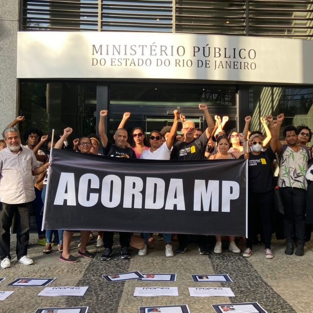 Fotografia mostra grupo de cerca de 30 pessoas com punho cerrado para cima segurando uma placa de fundo preto e letras brancas garrafais onde se lê "Acorda, MP". Elas estão na frente da entrada do Ministério Público do Estado do Rio de Janeiro, indicada pela placa acima delas. No chão em frente ao grupo, há cartazes com fotos de jovens vítimas da violência policial espalhados pelo chão.