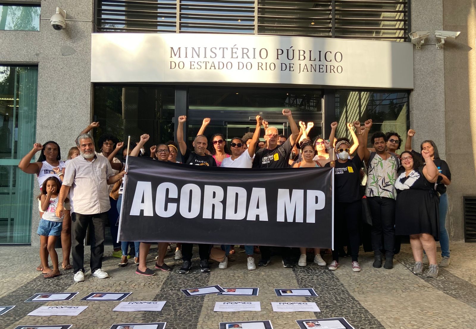 Fotografia mostra grupo de cerca de 30 pessoas com punho cerrado para cima segurando uma placa de fundo preto e letras brancas garrafais onde se lê "Acorda, MP". Elas estão na frente da entrada do Ministério Público do Estado do Rio de Janeiro, indicada pela placa acima delas. No chão em frente ao grupo, há cartazes com fotos de jovens vítimas da violência policial espalhados pelo chão.