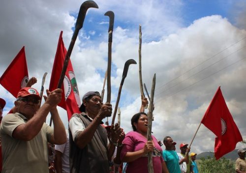 Grupo de trabalhadores rurais, homens e mulheres adultos, erguem foices e bandeiras vermelhas com o símbolo do MST em manifestação no campo. 